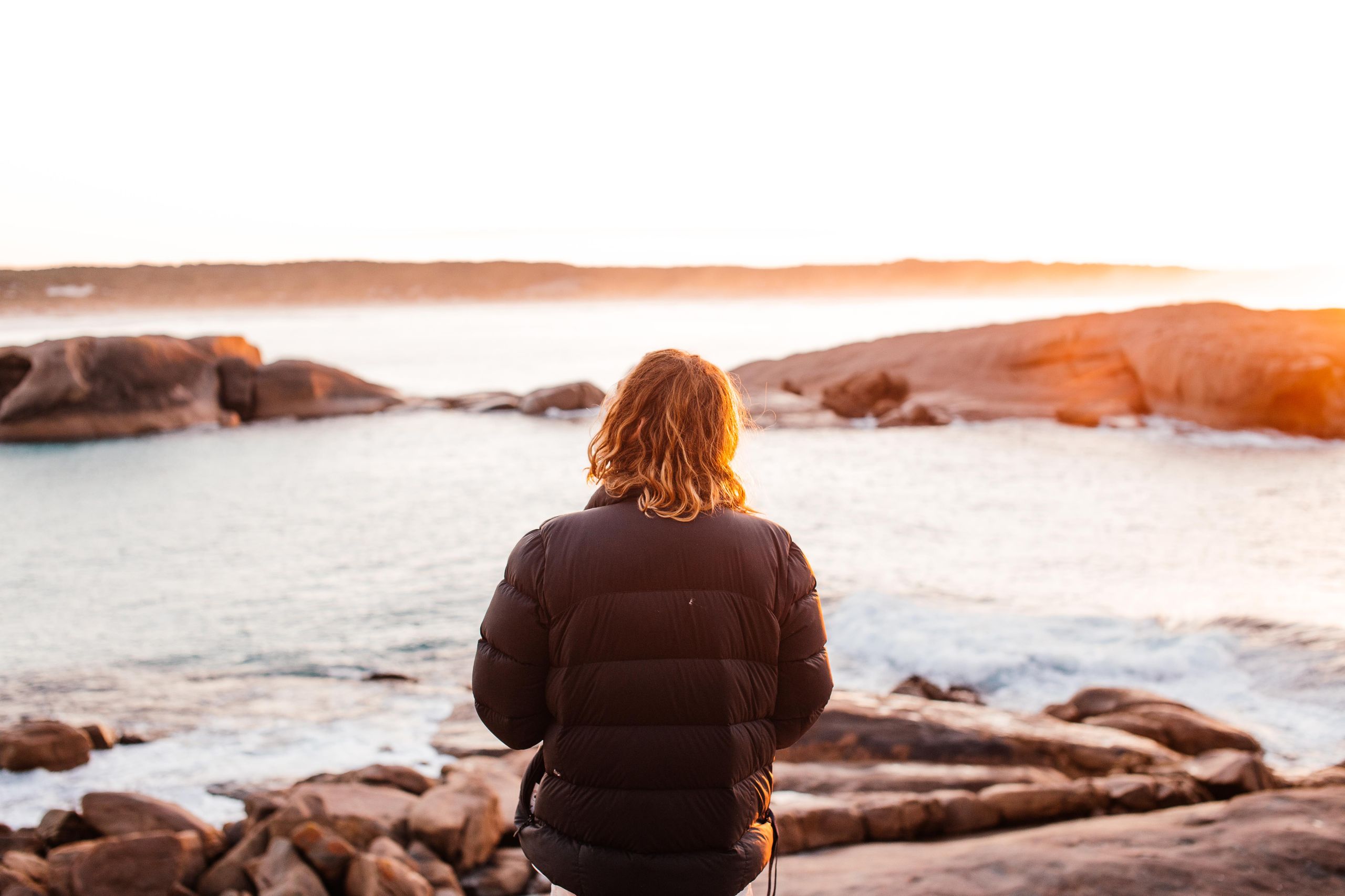 A person in a jacket views an ocean sunrise from rocky cliffs, capturing tranquility.