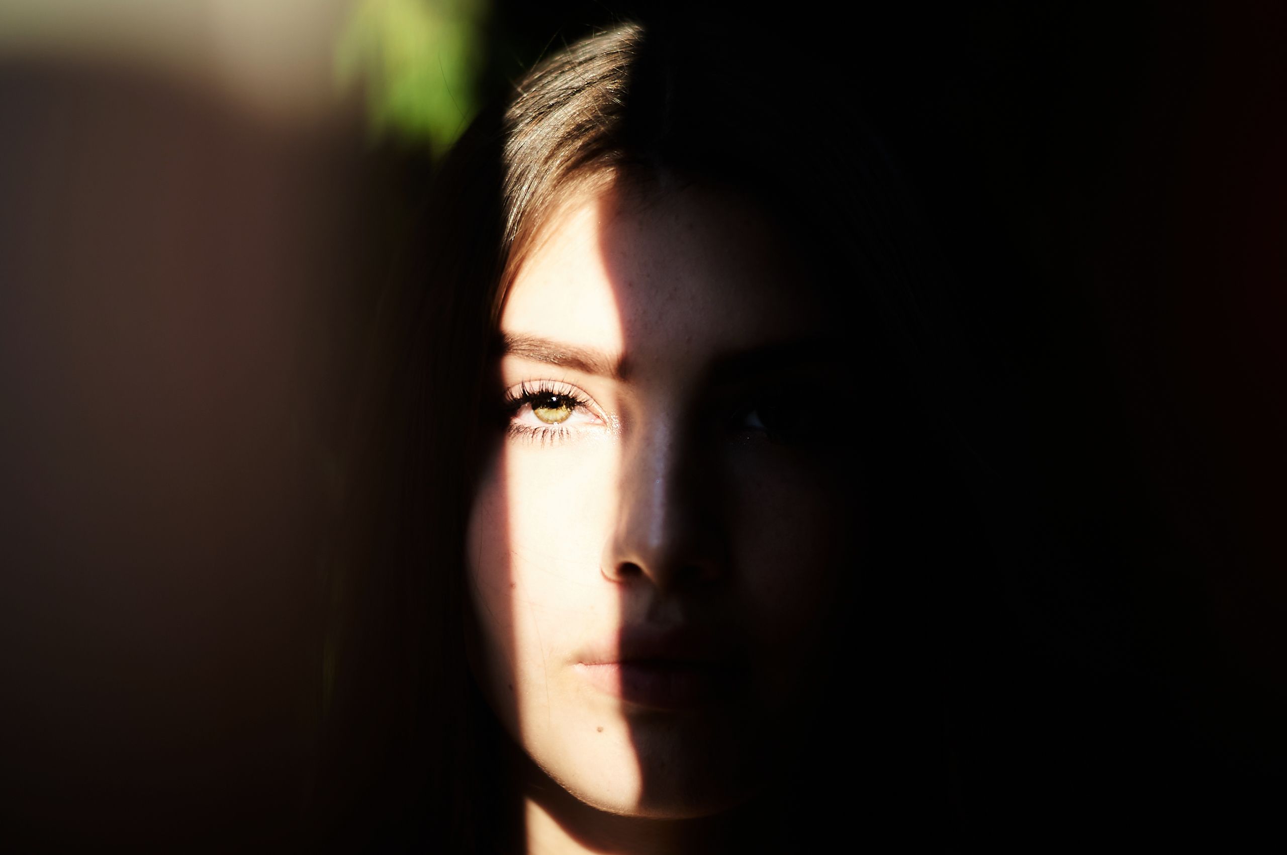 A woman’s face is partially illuminated by sunlight in a dark setting, creating a moody and dramatic effect.