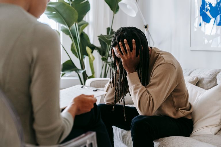 Anxious African American male with hands on head sitting on sofa near crop female psychologist with clipboard during appointment in office