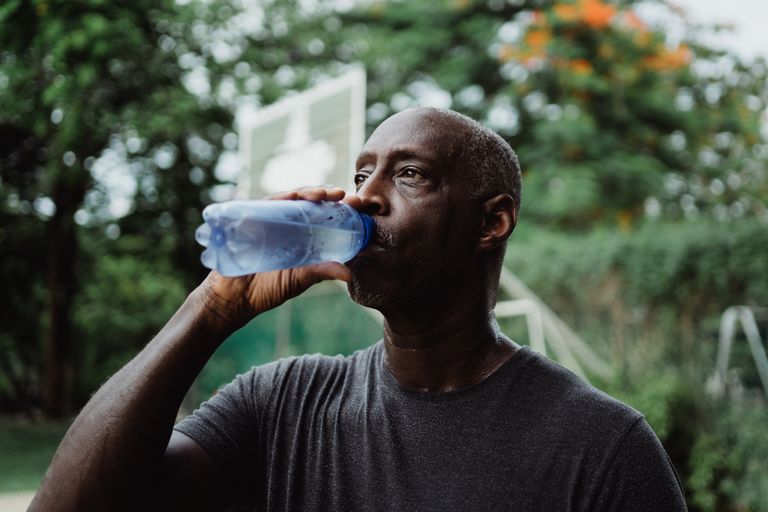 Mature African American man drinking water from a bottle in a park setting, staying hydrated.