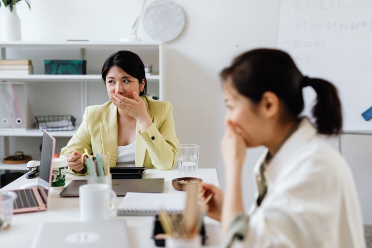 Two Asian women enjoying a light-hearted moment during a lunch break in an office setting.