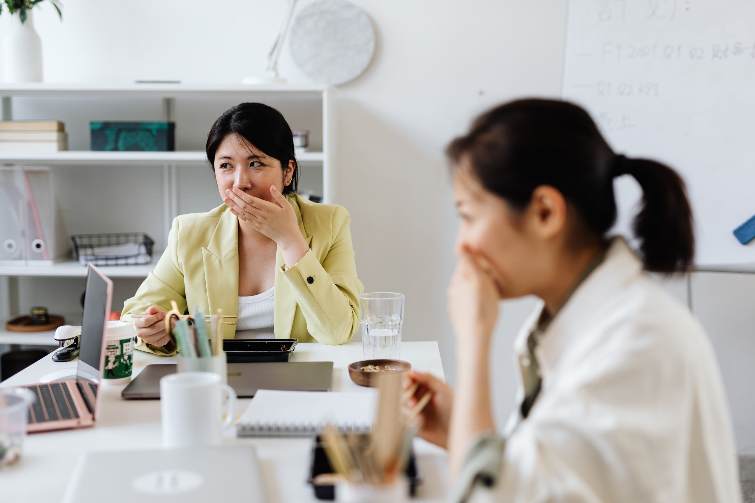 Two Asian women enjoying a light-hearted moment during a lunch break in an office setting.
